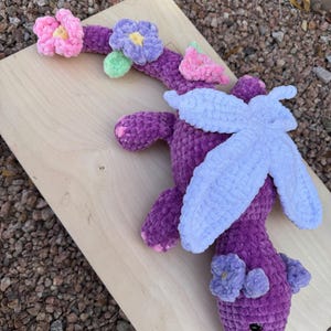 Puede incluir: Un drag&oacute;n de ganchillo morado hecho a mano con alas azul claro y detalles de flores rosas y moradas. El drag&oacute;n est&aacute; sobre una superficie de madera, mostrando su dise&ntilde;o suave y texturizado. La cabeza del drag&oacute;n tiene dos peque&ntilde;as flores moradas.