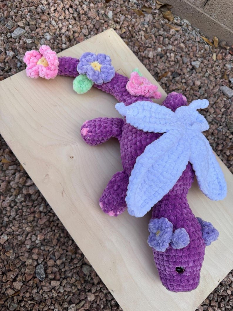 Puede incluir: Un drag&oacute;n de ganchillo morado hecho a mano con alas azul claro y detalles de flores rosas y moradas. El drag&oacute;n est&aacute; sobre una superficie de madera, mostrando su dise&ntilde;o suave y texturizado. La cabeza del drag&oacute;n tiene dos peque&ntilde;as flores moradas.