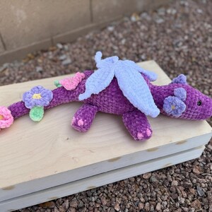Puede incluir: Un drag&oacute;n morado hecho a mano con ganchillo, con alas blancas y detalles de flores rosas y moradas. El drag&oacute;n est&aacute; tumbado sobre una superficie de madera de color claro. El cuerpo del drag&oacute;n es de color morado oscuro y las flores son rosas, moradas y verde claro.