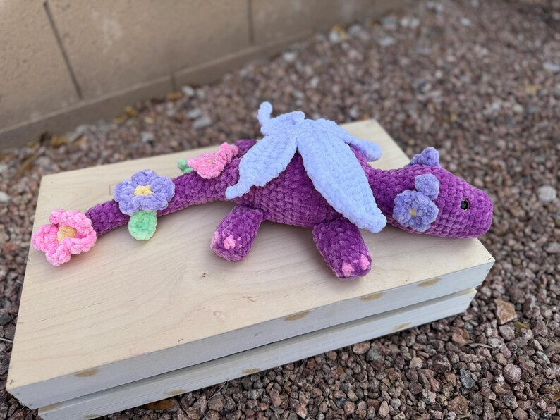 Puede incluir: Un drag&oacute;n morado hecho a mano con ganchillo, con alas blancas y detalles de flores rosas y moradas. El drag&oacute;n est&aacute; tumbado sobre una superficie de madera de color claro. El cuerpo del drag&oacute;n es de color morado oscuro y las flores son rosas, moradas y verde claro.