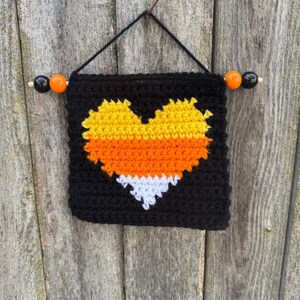 May include: A black, crocheted wall hanging with a heart-shaped design in the colors of candy corn: yellow, orange, and white. The hanging is suspended by a black string and wooden dowel with orange and black beads.
