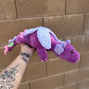 Puede incluir: Un drag&oacute;n de ganchillo hecho a mano en tonos morado, rosa y blanco. El drag&oacute;n presenta peque&ntilde;os detalles de flores y alas. El peluche se sostiene contra un fondo de pared de ladrillo.