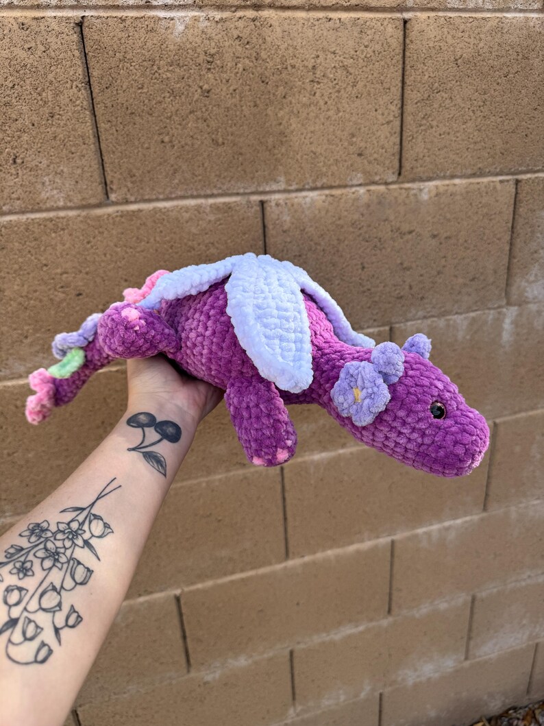Puede incluir: Un drag&oacute;n de ganchillo hecho a mano en tonos morado, rosa y blanco. El drag&oacute;n presenta peque&ntilde;os detalles de flores y alas. El peluche se sostiene contra un fondo de pared de ladrillo.