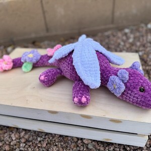 Puede incluir: Un drag&oacute;n de ganchillo morado hecho a mano con alas azul claro y detalles de flores rosas y moradas. El drag&oacute;n est&aacute; tumbado sobre una caja de madera de color claro, mostrando su dise&ntilde;o suave y texturizado. Los ojos del drag&oacute;n son negros.