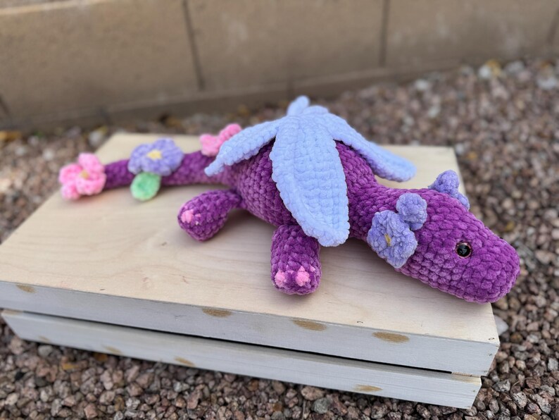 Puede incluir: Un drag&oacute;n de ganchillo morado hecho a mano con alas azul claro y detalles de flores rosas y moradas. El drag&oacute;n est&aacute; tumbado sobre una caja de madera de color claro, mostrando su dise&ntilde;o suave y texturizado. Los ojos del drag&oacute;n son negros.