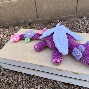 Puede incluir: Un drag&oacute;n de ganchillo morado hecho a mano con alas azul claro y detalles de flores. El drag&oacute;n est&aacute; sobre una superficie de madera clara. El cuerpo del drag&oacute;n es de color p&uacute;rpura oscuro, con detalles de flores rosas y moradas claras.