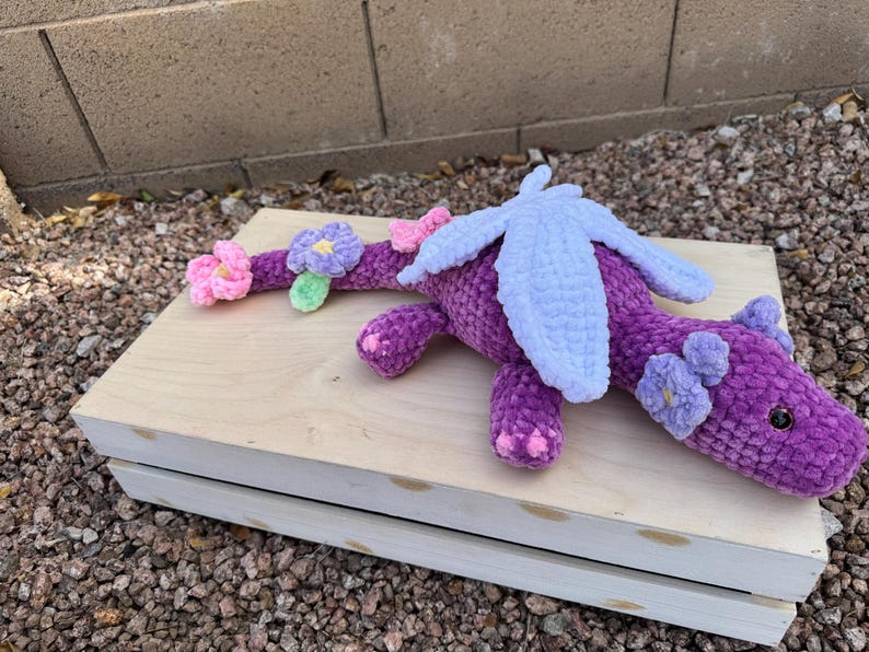 Puede incluir: Un drag&oacute;n de ganchillo morado hecho a mano con alas azul claro y detalles de flores. El drag&oacute;n est&aacute; sobre una superficie de madera clara. El cuerpo del drag&oacute;n es de color p&uacute;rpura oscuro, con detalles de flores rosas y moradas claras.