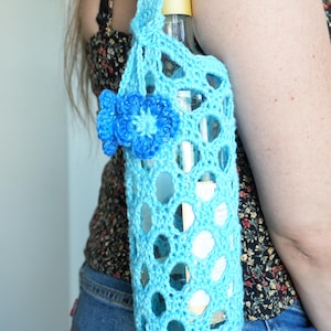 May include: A light blue crocheted bottle holder featuring an openwork mesh pattern and a decorative blue flower accent. The holder includes a single handle and is designed to carry a glass bottle with a gold-colored cap, showcasing a textured yarn construction.