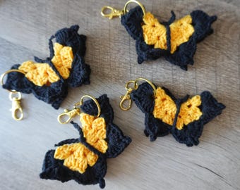 Puede incluir: Cuatro llaveros de mariposa tejidos a mano con hilo negro y amarillo. Cada mariposa está unida a un cierre de mosquetón de metal dorado y a una anilla circular, mostrando una textura de punto detallada y un contraste de colores vivo.