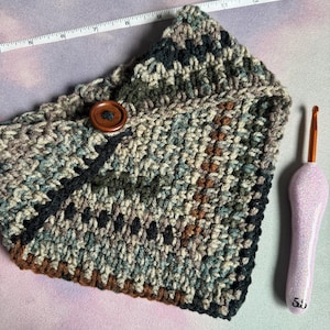 May include: A crocheted neck warmer in a camouflage pattern with brown, black, and green yarn. The warmer has a wooden button and is lying on a pink and purple surface.