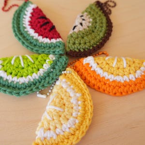 May include: Five crocheted fruit slice keychains arranged in a circular pattern. The set includes a red watermelon slice, a green kiwi slice with brown edges, a lime slice, an orange slice, and a yellow lemon slice, each with a metal ball chain.