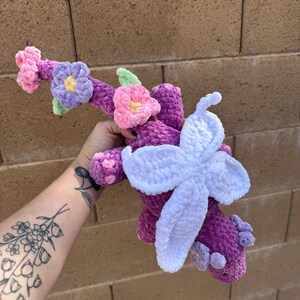 Puede incluir: Un peluche de ganchillo hecho a mano en tonos morado, rosa y blanco, adornado con detalles florales. El juguete presenta una gran flor de color morado claro y flores rosas y moradas m&aacute;s peque&ntilde;as. El peluche se sostiene contra una pared de ladrillos.