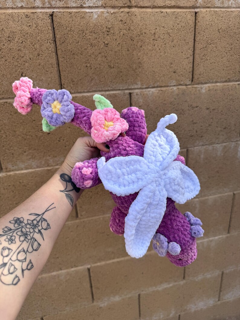 Puede incluir: Un peluche de ganchillo hecho a mano en tonos morado, rosa y blanco, adornado con detalles florales. El juguete presenta una gran flor de color morado claro y flores rosas y moradas m&aacute;s peque&ntilde;as. El peluche se sostiene contra una pared de ladrillos.