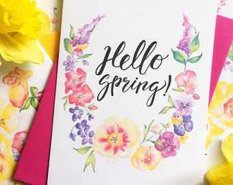 May include: A white card with a watercolor floral wreath and the text "Hello Spring!" in black calligraphy. The card is surrounded by yellow daffodils.