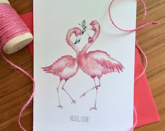 May include: A greeting card with a watercolor illustration of two pink flamingos forming a heart shape with their necks. The card reads "HELLO, LOVE!" and is accompanied by a pink envelope and twine.