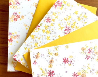 May include: A set of four note cards with a white background and a floral pattern in shades of yellow, pink, and orange. The cards are layered on top of each other, with the top card being a solid yellow color.
