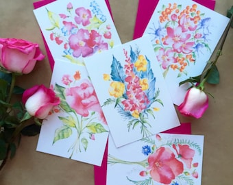 May include: Five watercolor paintings on white cardstock featuring different floral bouquets. The bouquets are painted in shades of pink, yellow, blue, and green. The cards are displayed on a brown surface.