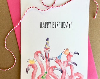 May include: A white greeting card with the text "HAPPY BIRTHDAY!" features a watercolor illustration of pink flamingos wearing party hats and crowns. A pink envelope and red and white twine complete the design.