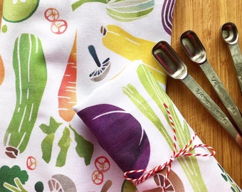 May include: White kitchen towels with colorful vegetable illustrations, including carrots, zucchini, and mushrooms. The towels are tied with red and white twine. Stainless steel measuring spoons are on a wooden surface.