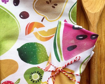 May include: White kitchen towel with colorful fruit illustrations, including watermelon, kiwi, and pear. A rolled-up napkin with a watermelon slice design is tied with red and white twine. A wooden spoon rests on a wooden surface.