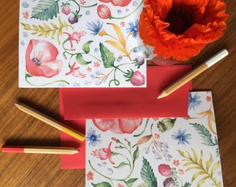 May include: Three cards with a floral pattern featuring red poppies, green leaves, and white flowers. The cards are on a wooden surface with two colored pencils.