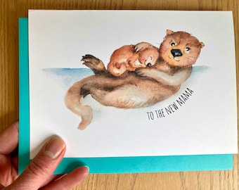 May include: Greeting card featuring a watercolor illustration of two otters. One otter is cradling a smaller otter. The card has the text "TO THE NEW MAMA" in black script. The card is on a teal envelope.