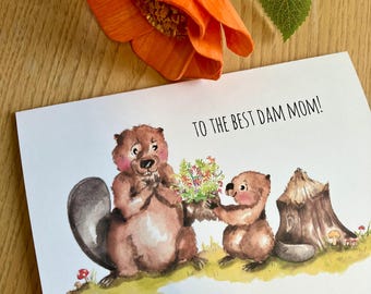 May include: A white card with a watercolor illustration of two beavers. The beaver on the left is holding a bouquet of flowers and the beaver on the right is holding a single flower. The card says "To the best dam mom!" in black text.