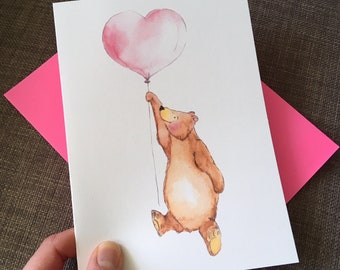 May include: A watercolor illustration of a brown bear holding a pink heart-shaped balloon. The bear is depicted with a cheerful expression, and the balloon is attached by a thin string. A pink envelope is behind the card.