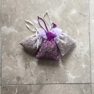 May include: Three small lavender sachets, two are white with purple trim and one is purple with white trim. The sachets are filled with dried lavender and are tied with white ribbon.