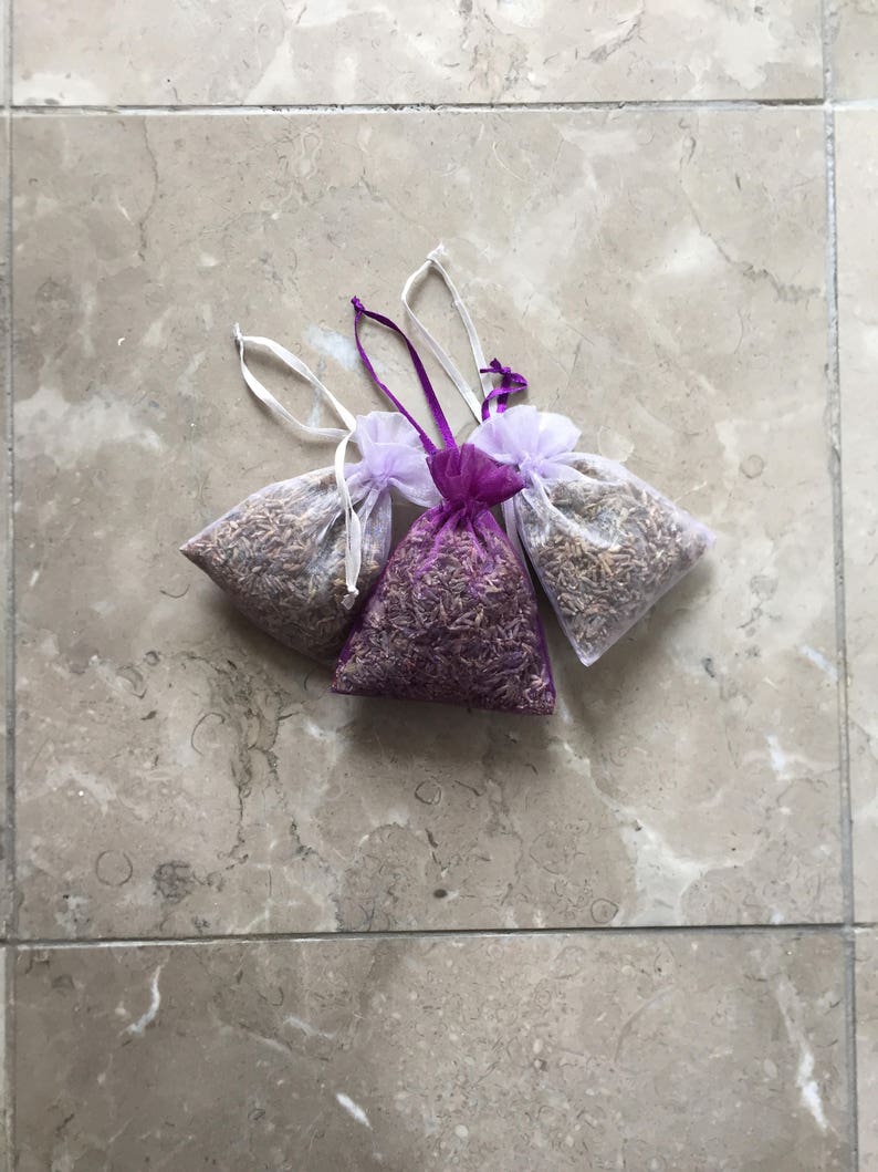 May include: Three small lavender sachets, two are white with purple trim and one is purple with white trim. The sachets are filled with dried lavender and are tied with white ribbon.