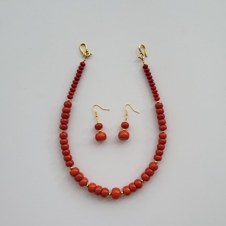 Op de afbeelding: Een rode koraal kralenketting en bijpassende oorbellen. De ketting heeft een gouden sluiting en de oorbellen hebben gouden haakjes.