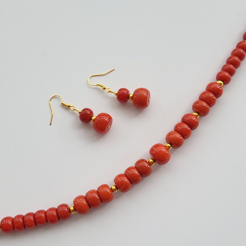 Op de afbeelding: Een rode en gouden kralenketting en bijpassende oorbellen. De ketting is gemaakt van ronde, rode kralen met gouden tussenstukken. De oorbellen zijn gemaakt van twee rode kralen met een gouden haak.
