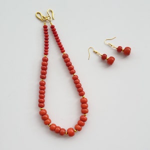 Op de afbeelding: Een rode koraal kralenketting en bijpassende oorbellen. De ketting is gemaakt van kleine, ronde kralen en heeft een gouden sluiting. De oorbellen zijn ook gemaakt van kleine, ronde kralen en hebben gouden haakjes.
