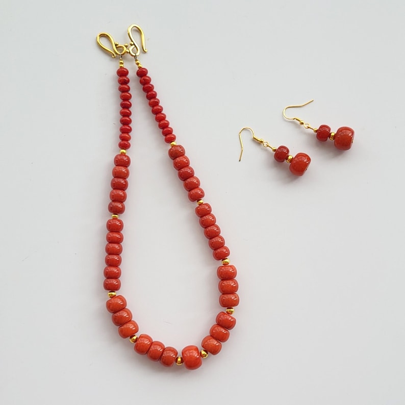 Op de afbeelding: Een rode koraal kralenketting en bijpassende oorbellen. De ketting is gemaakt van kleine, ronde kralen en heeft een gouden sluiting. De oorbellen zijn ook gemaakt van kleine, ronde kralen en hebben gouden haakjes.