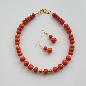 May include: A red beaded necklace with gold accents and matching earrings. The necklace is made of round beads and has a gold clasp. The earrings are also made of round beads and have gold hooks.