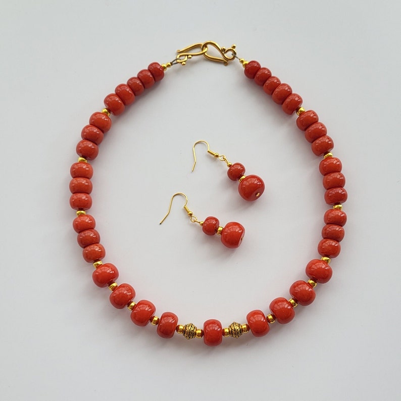 May include: A red beaded necklace with gold accents and matching earrings. The necklace is made of round beads and has a gold clasp. The earrings are also made of round beads and have gold hooks.