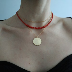 May include: A layered necklace with a gold pendant. The necklace features a red beaded chain and two gold chains. The pendant is a large, round, gold disc.