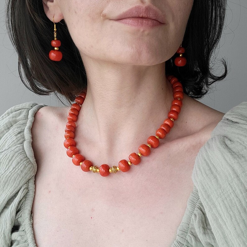 May include: A coral necklace with gold accents. The necklace is made of round, orange coral beads. The necklace is worn by a person with a light green shirt.