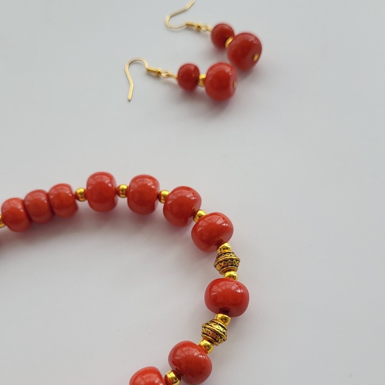 May include: A red beaded necklace with gold accents. The necklace is made of round beads and has a simple design. The necklace is paired with matching earrings.