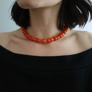 May include: A close-up of a red coral bead necklace with gold accents. The necklace is worn by a person with short dark hair.