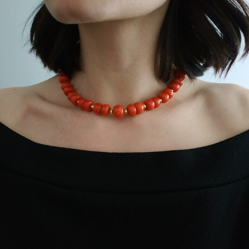 May include: A close-up of a red coral bead necklace with gold accents. The necklace is worn by a person with short dark hair.