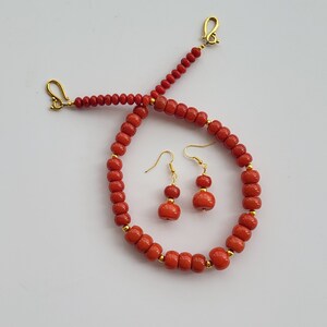Op de afbeelding: Een rode koraal ketting en oorbellen set met gouden accenten. De ketting is gemaakt van ronde koraal kralen en heeft een gouden sluiting. De oorbellen zijn hangende oorbellen met twee ronde koraal kralen en gouden haken.
