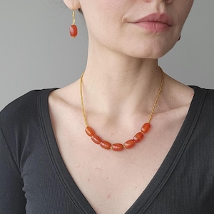 May include: A gold necklace with a row of orange carnelian beads. The necklace is worn by a person with a pair of matching carnelian earrings.
