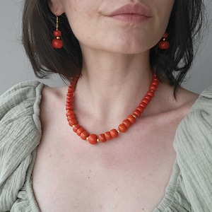 Op de afbeelding: Een rode koraal ketting met gouden accenten. De ketting is gemaakt van ronde kralen en wordt gedragen door een persoon met donker haar.