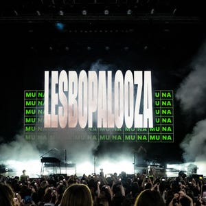 May include: A live concert scene with a large screen displaying the text "LESBOPALOOZA" in bold white letters. The stage is lit with bright lights, and a crowd of people is visible in the foreground, many holding up phones.