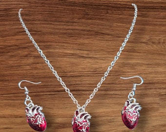 May include: A silver-toned necklace and earring set featuring anatomical heart pendants. The heart pendants are a deep red color with silver accents, and the necklace and earrings have a delicate chain design. The set is displayed on a wooden surface.