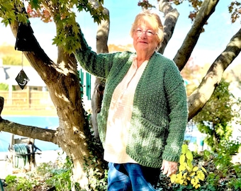 Pode incluir: Uma mulher vestindo um casaco de tricô verde com um bolso, uma camisa rosa claro e calças jeans azuis está em pé na frente de uma árvore.