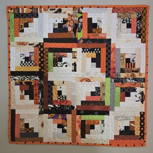 May include: A handmade Halloween-themed quilt with a patchwork design. The quilt features a variety of fabrics in orange, black, white, and green. The quilt has an orange border with black polka dots and is hanging on a gray wall.