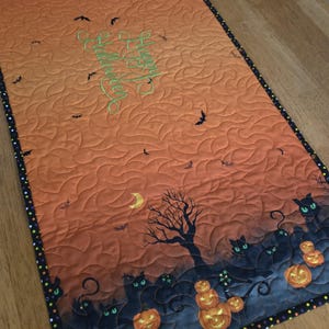 May include: Halloween-themed quilted table runner with an orange and black gradient background. Features embroidered text, bats, a crescent moon, and silhouettes of cats, pumpkins, and a bare tree. The border is black with colorful polka dots.