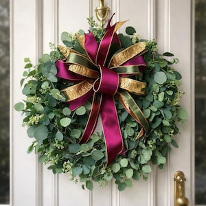 May include: A round eucalyptus wreath with a large burgundy and gold ribbon bow. The wreath is adorned with small white flowers and is hanging on a white door with gold hardware. The bow has a gold patterned trim.